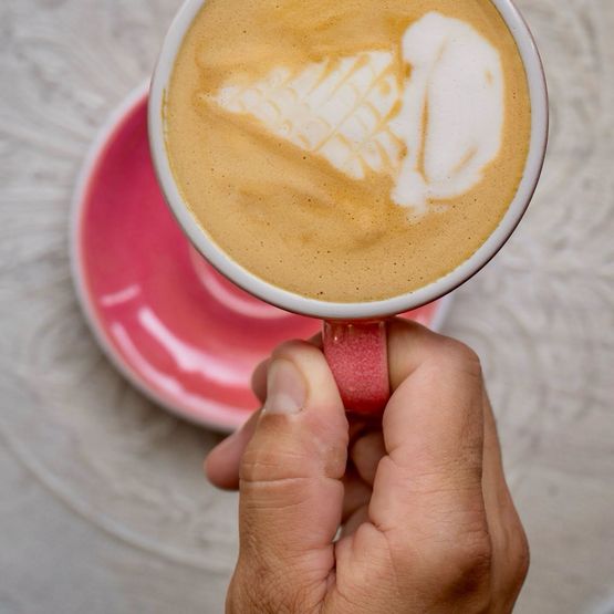Ruka držící růžový šálek kávy s latte artem ve tvaru zmrzlinového kornoutu.